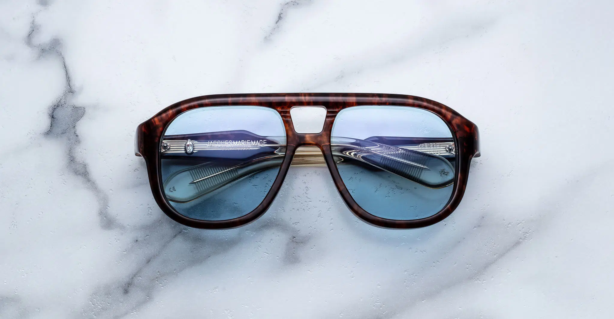 Jacques-Marie-Mage-Jasper-Argyle-JMMYA-5C-Front-Dan-Deutsch A pair of Jacques Marie Mage Jasper tortoiseshell sunglasses with blue-tinted lenses rests on white marble, reflecting a folded pair of silver-framed eyeglasses in their lenses.