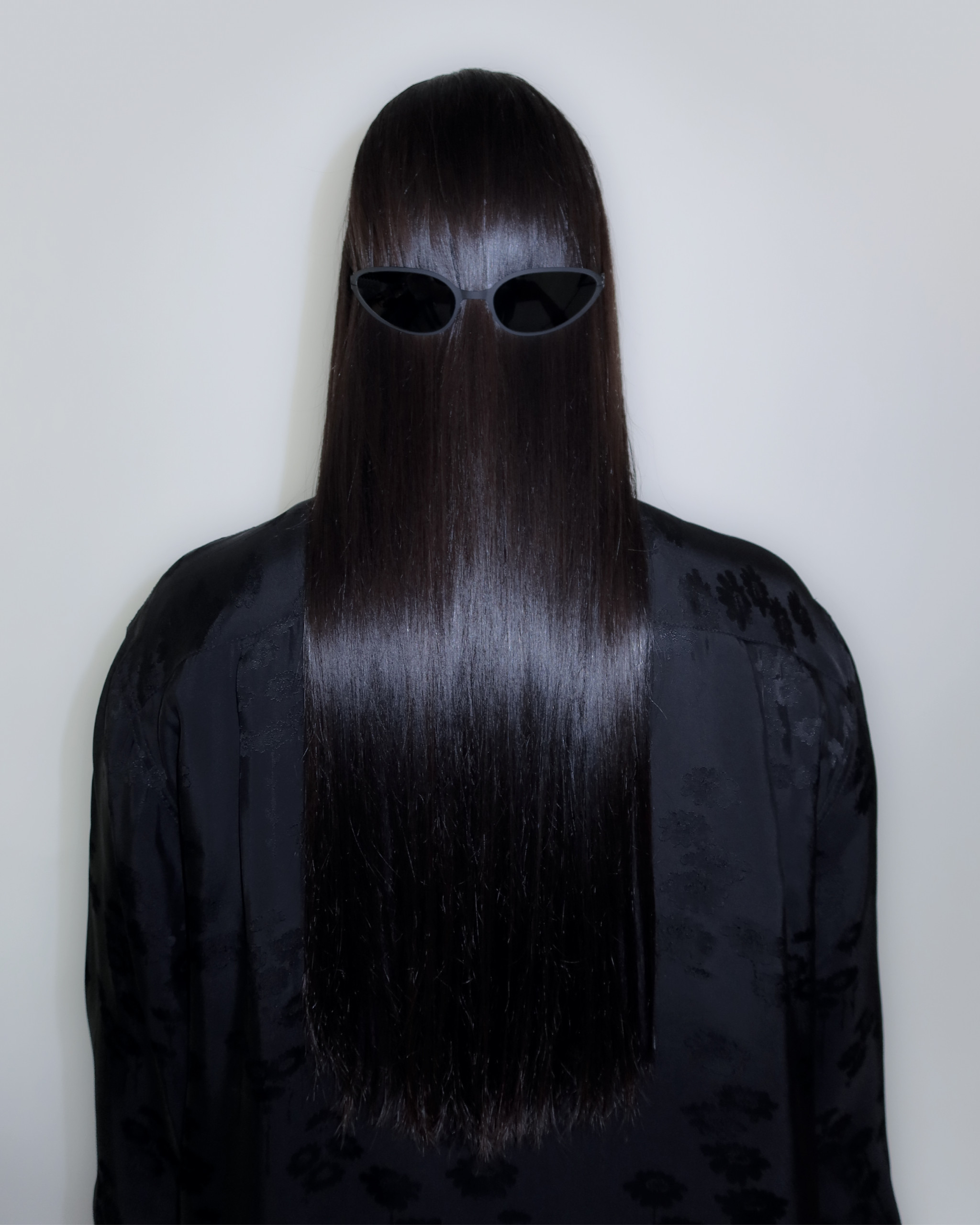 A person with long, straight, dark hair faces away from the camera, wearing black cat-eye Mykita sunglasses on the back of their head, creating a quirky and surreal effect. They are dressed in a black outfit against a plain background.
