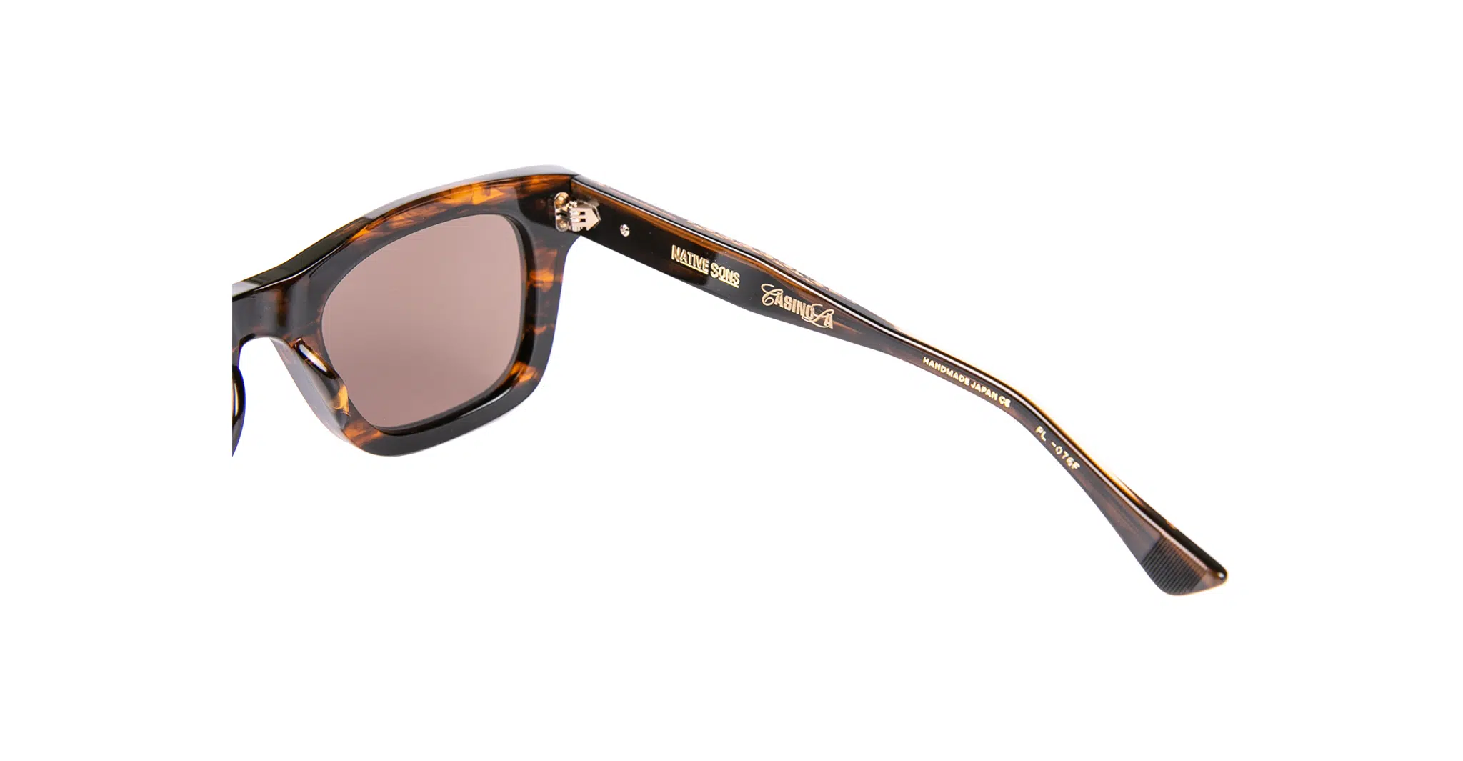 Close-up of the arm of Native Sons x Casinola "The E's" brown tortoiseshell sunglasses featuring a gold logo and printed text inside. The lens is tinted, set against a white background.