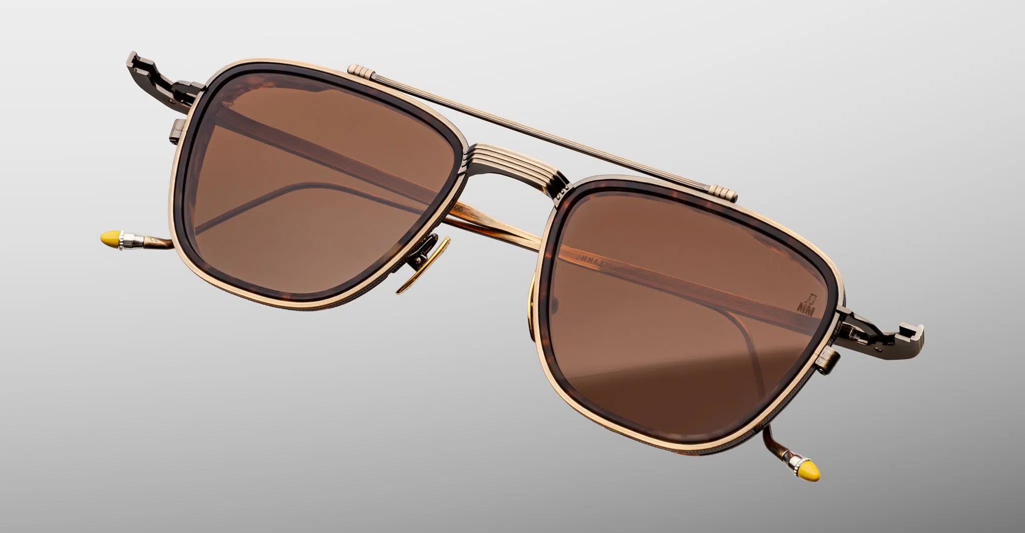 The Jacques Marie Mage 1992 are stylish gold-framed sunglasses with large brown-tinted lenses, a double bridge, decorative gold accents, and yellow temple tips set against a light gray background.