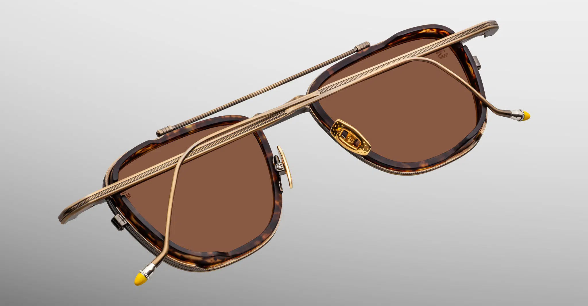 Close-up of Jacques Marie Mage 1992 sunglasses with brown lenses, tortoiseshell frames, and gold-toned metal arms featuring yellow accents at the tips, set against a light grey background.