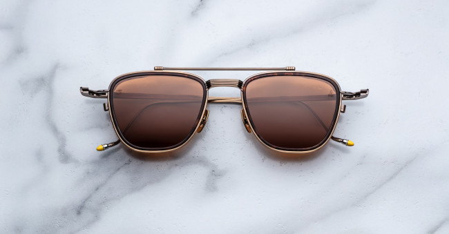 A pair of Jacques Marie Mage 1992 sunglasses, featuring brown-tinted square aviator lenses and a metal frame, is displayed on a white marble surface.
