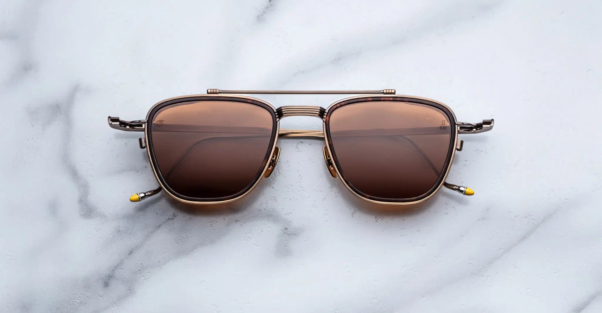 A pair of Jacques Marie Mage 1992 sunglasses, featuring brown-tinted square aviator lenses and a metal frame, is displayed on a white marble surface.