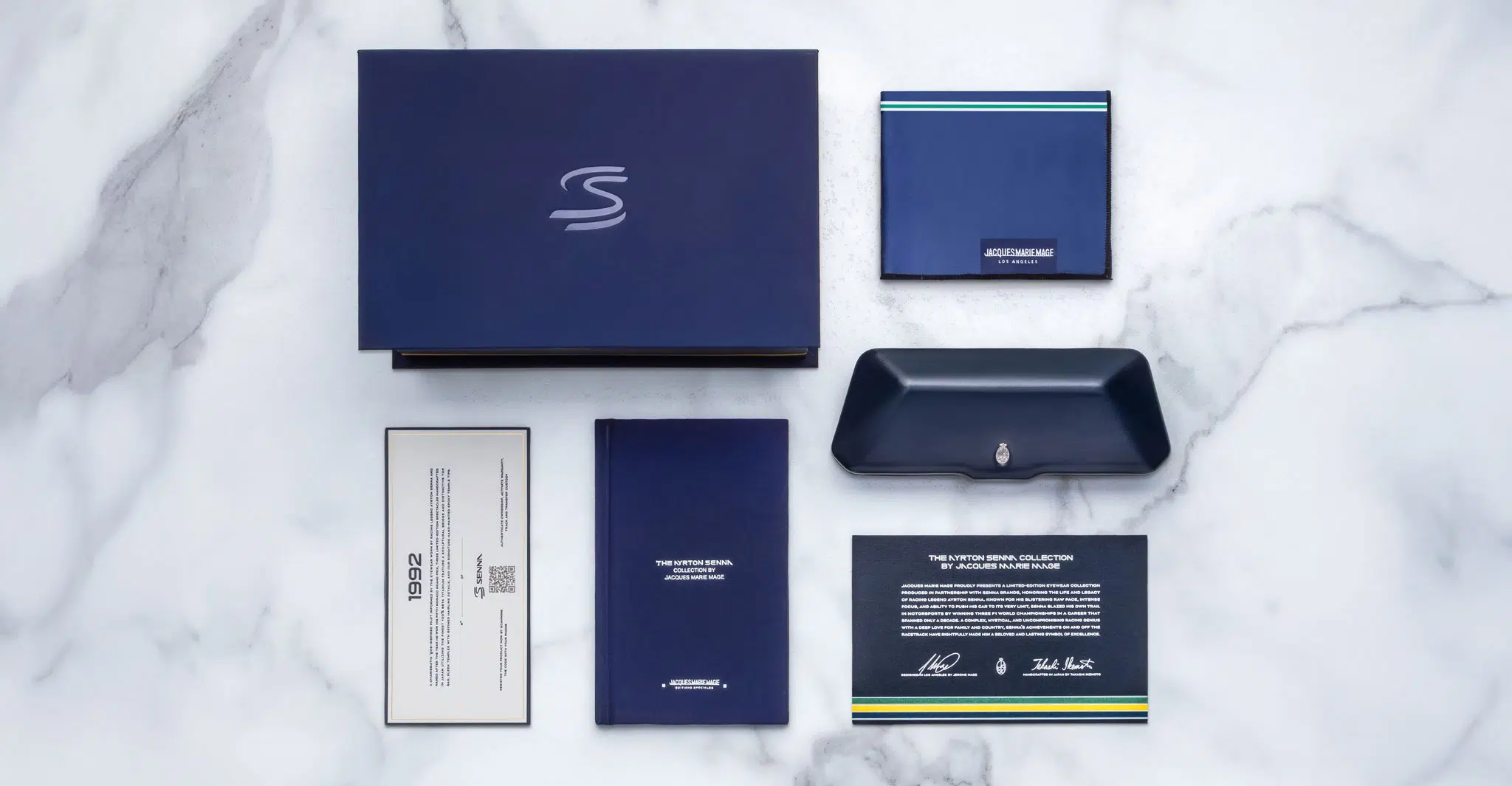 A flat lay of the Jacques Marie Mage 1992 set—dark blue box, matching wallet, pen case, booklet, certificate, and info card with silver logos—all arranged neatly on white marble.