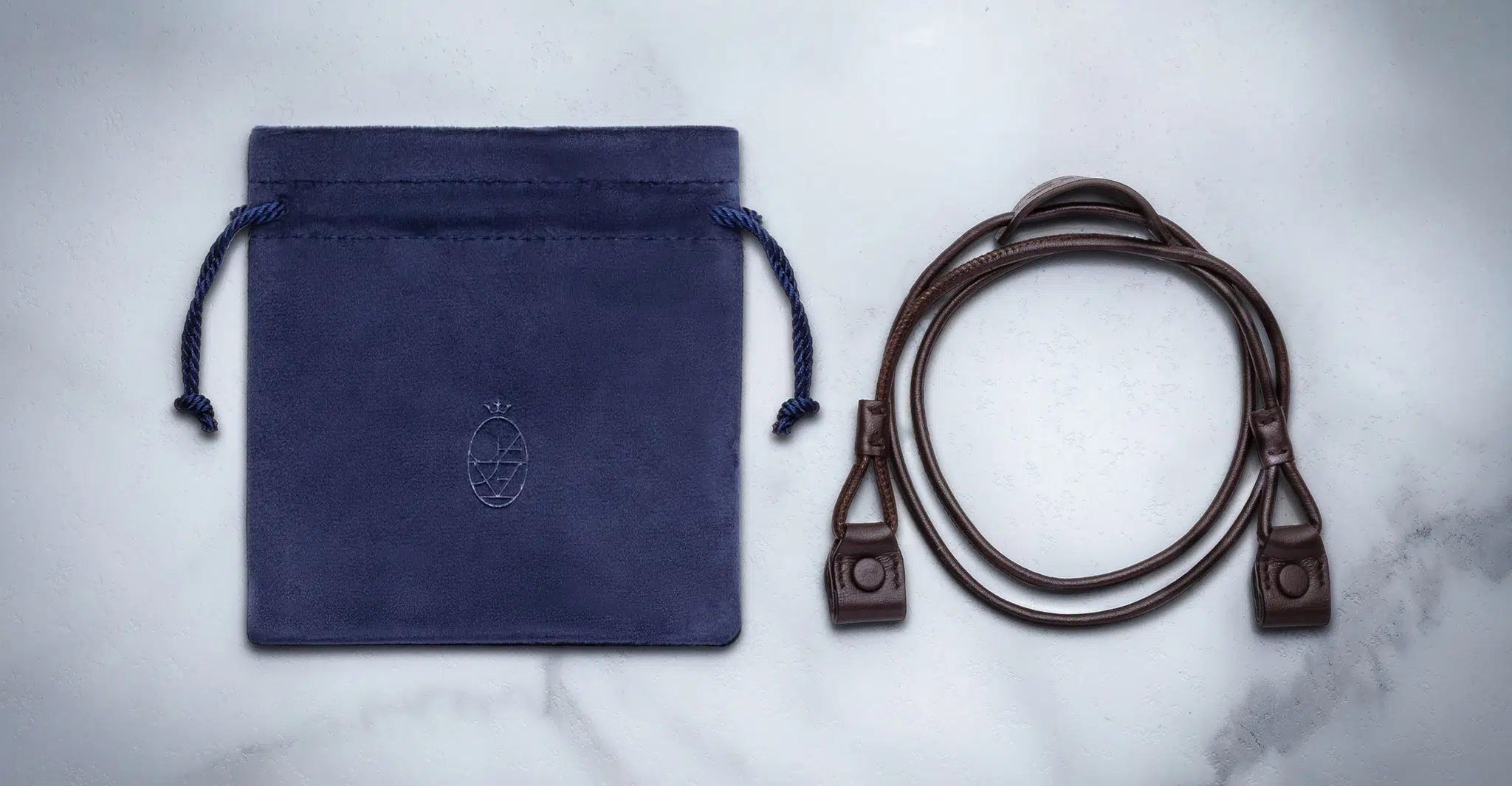 The Jacques Marie Mage 1992, featuring a blue suede drawstring pouch with embossed logo and a coiled dark brown leather strap with clasps, is displayed on a light marble surface.