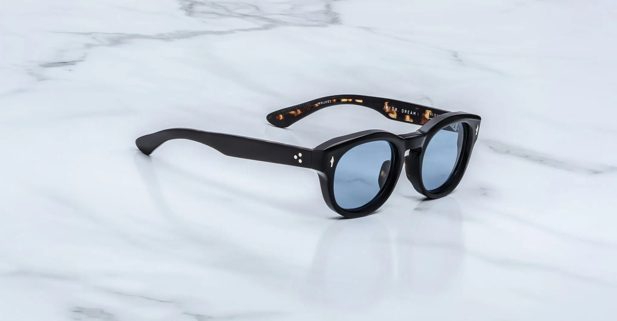 Jacques-Marie-Mage-Dr-Dream-Noir-JMMJH-01-3-4-Dan-Deutsch The Jacques Marie Mage Dr. Dream sunglasses feature black round frames with blue-tinted lenses, resting on a white marble surface. The inner arms display a tortoiseshell pattern accented by gold text.