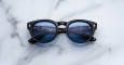 A pair of Jacques Marie Mage Dr. Dream round black sunglasses with blue-tinted lenses rests on a white marble surface, reflecting text from a computer screen.