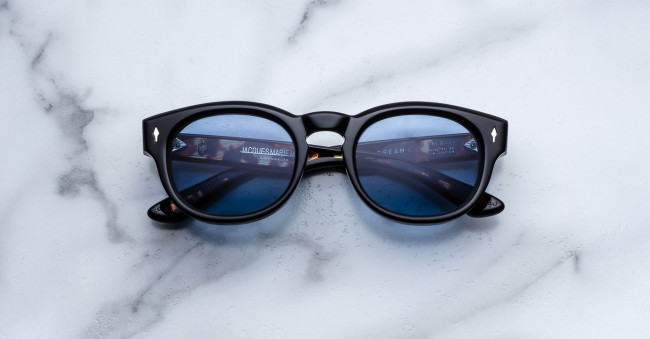 A pair of Jacques Marie Mage Dr. Dream round black sunglasses with blue-tinted lenses rests on a white marble surface, reflecting text from a computer screen.