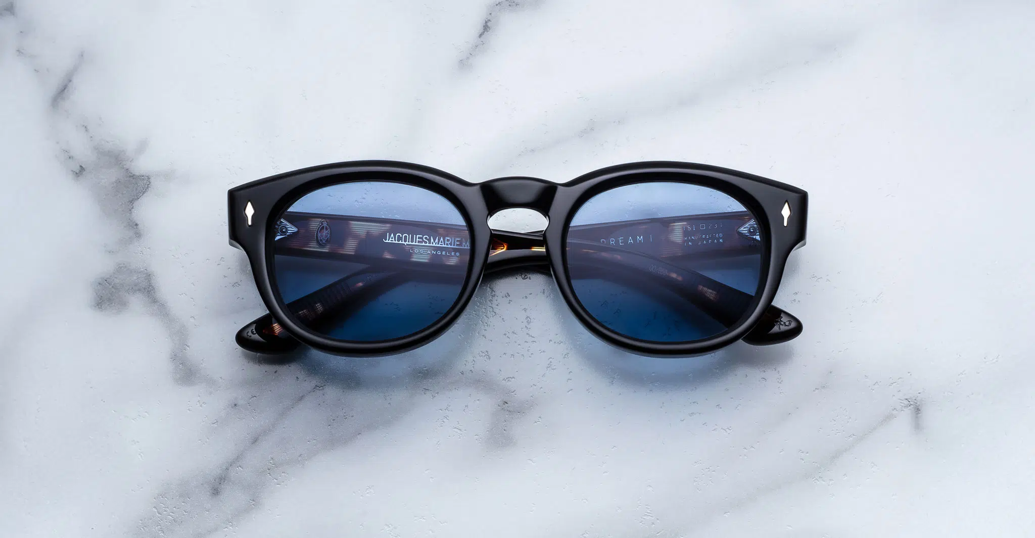 Jacques-Marie-Mage-Dr-Dream-Noir-JMMJH-01-Front-Dan-Deutsch A pair of Jacques Marie Mage Dr. Dream round black sunglasses with blue-tinted lenses rests on a white marble surface, reflecting text from a computer screen.
