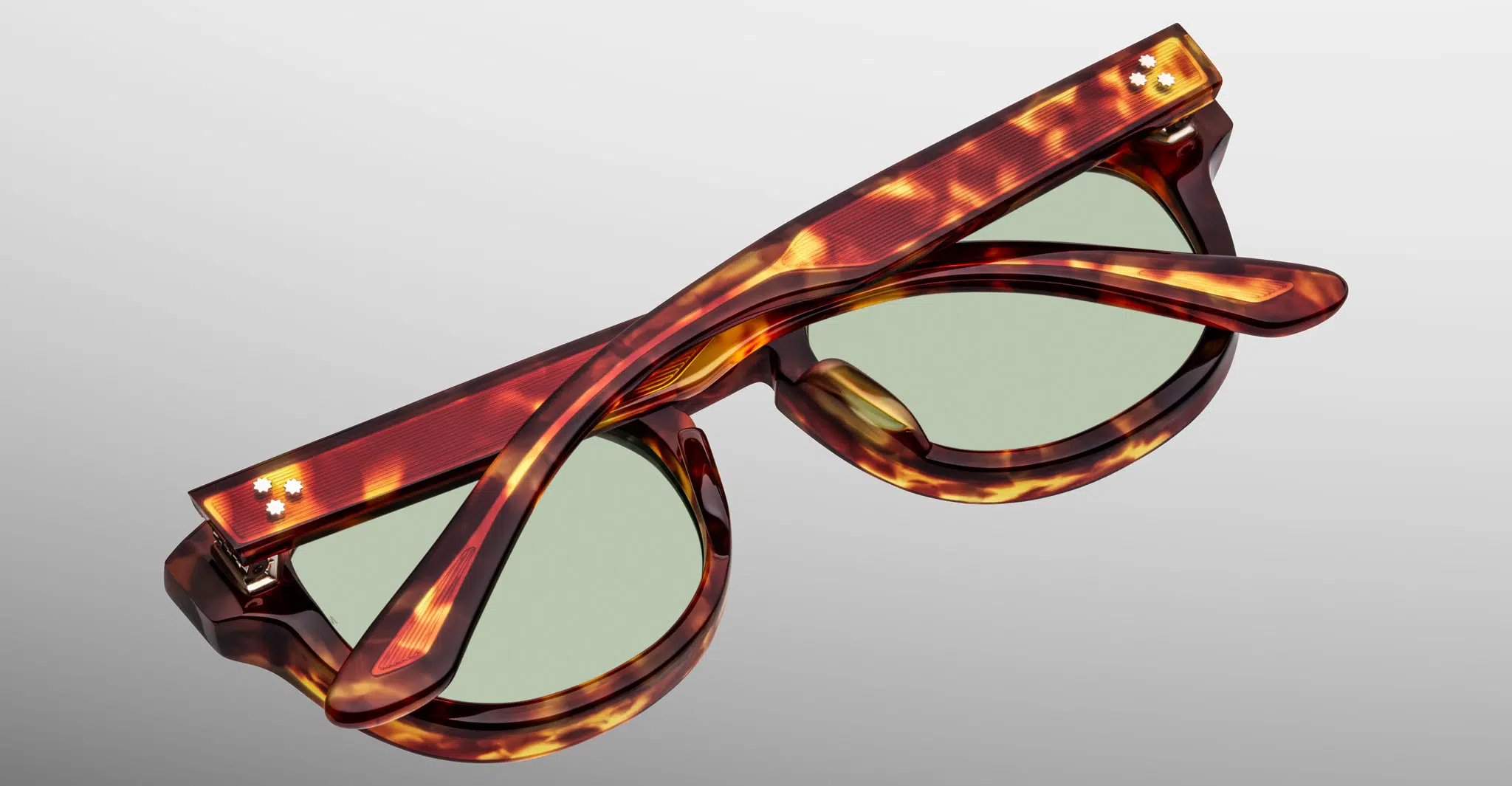 The Jacques Marie Mage Dr. Dream sunglasses feature a tortoiseshell pattern and green lenses, displayed folded with crossed arms on a plain light gray background.