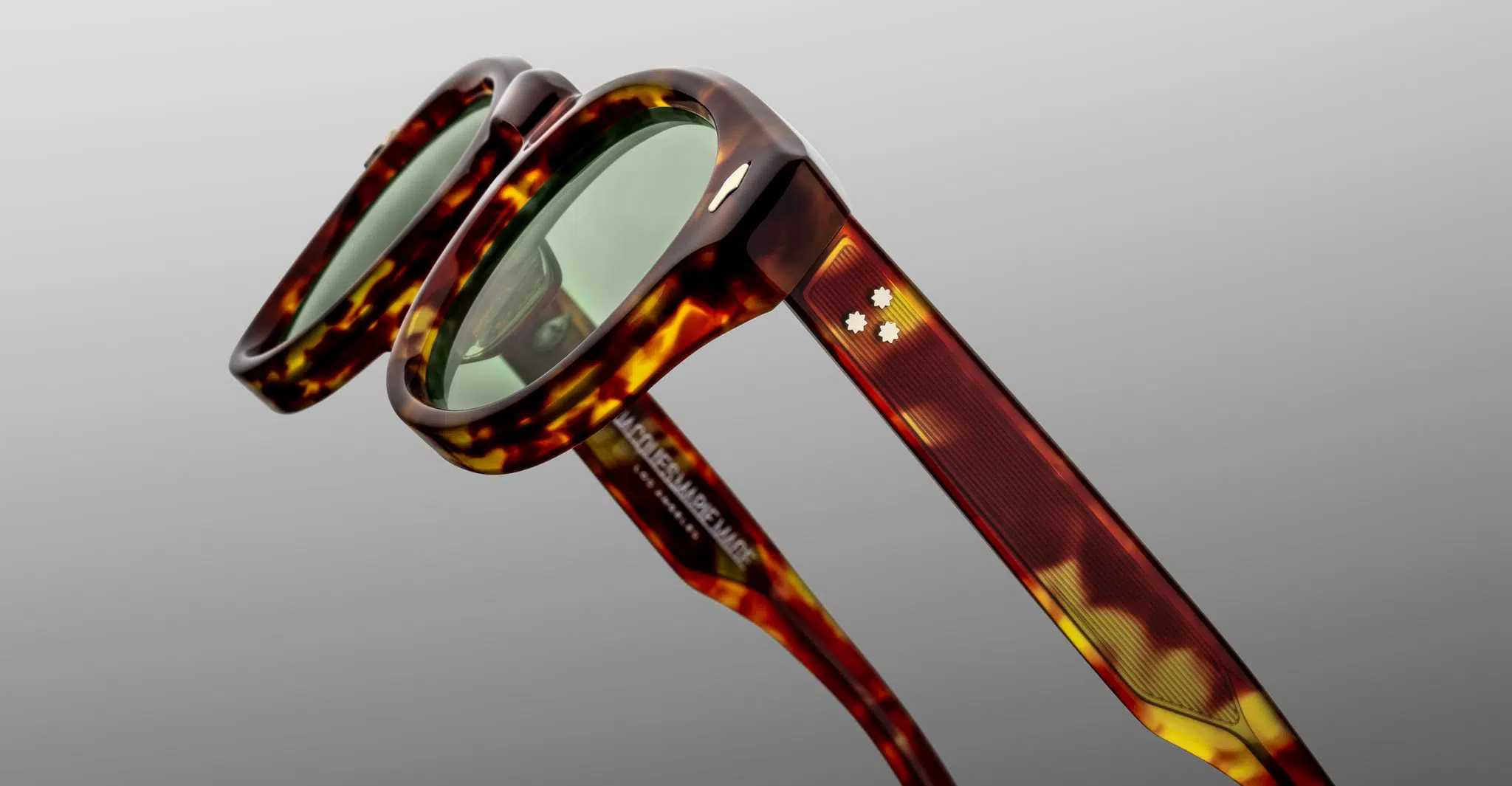 Close-up of Jacques Marie Mage Dr. Dream sunglasses, featuring green lenses, a textured tortoiseshell frame in brown and yellow, and two small white star-shaped accents on the temples against a smooth gray background.