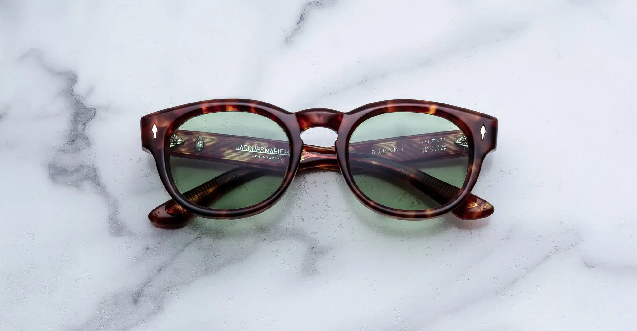A pair of Jacques Marie Mage Dr. Dream tortoiseshell sunglasses with green lenses rests on a white marble surface. The arms are crossed, and light reflections shine on the lenses.