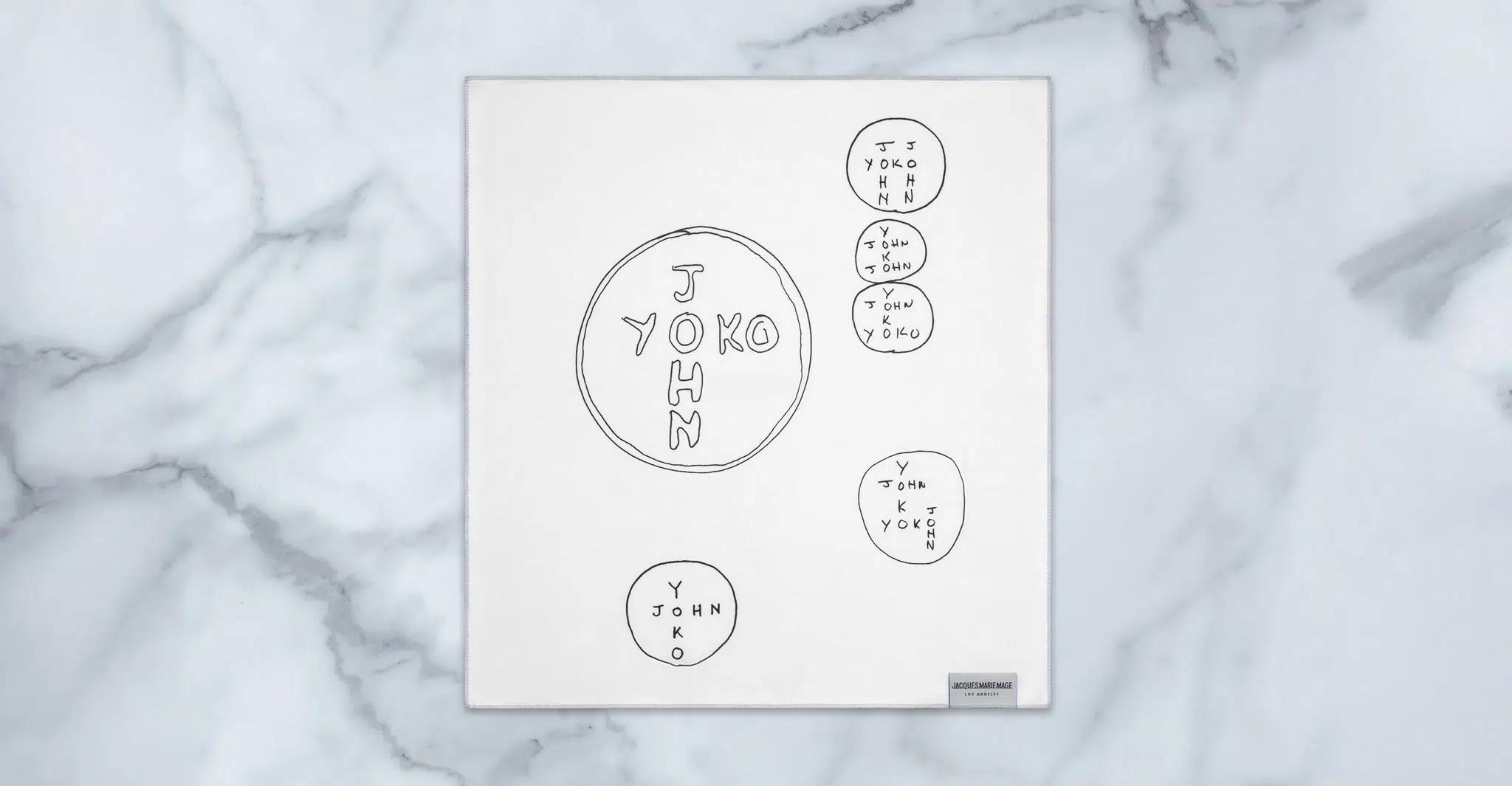A white square cloth on marble displays hand-drawn circles and overlapping text like "YOKO," "JOHN," and "YOKO + JOHN" in black ink, inspired by the Jacques Marie Mage Ocean Child collection.