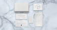 A flat lay of Jacques Marie Mage Ocean Child luxury packaging on marble, featuring a white box, round-labeled pouch, drawstring bag, info cards, and small envelope&mdash;all with minimalist designs and embossed or printed text.