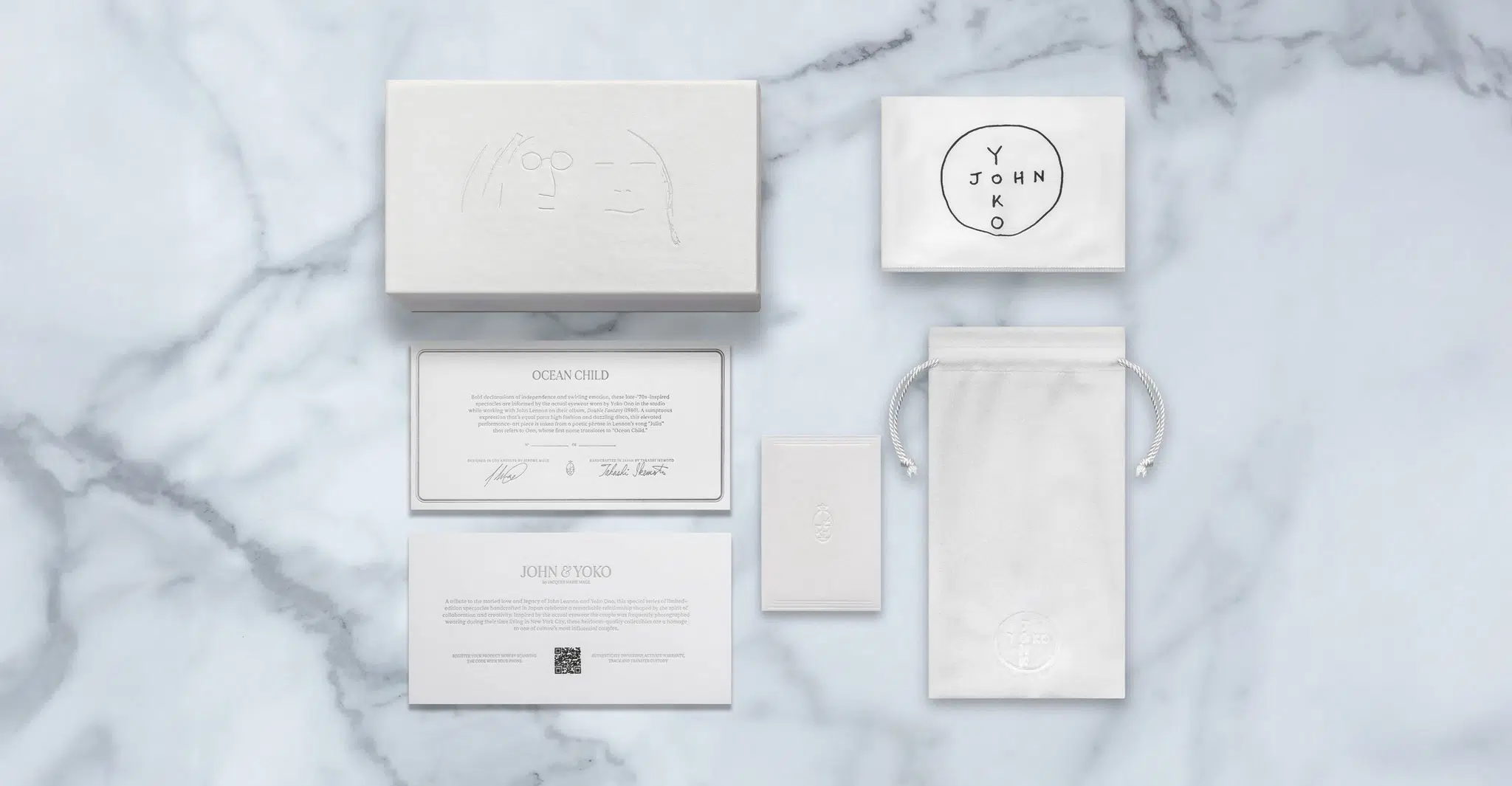 A flat lay of Jacques Marie Mage Ocean Child luxury packaging on marble, featuring a white box, round-labeled pouch, drawstring bag, info cards, and small envelope&mdash;all with minimalist designs and embossed or printed text.