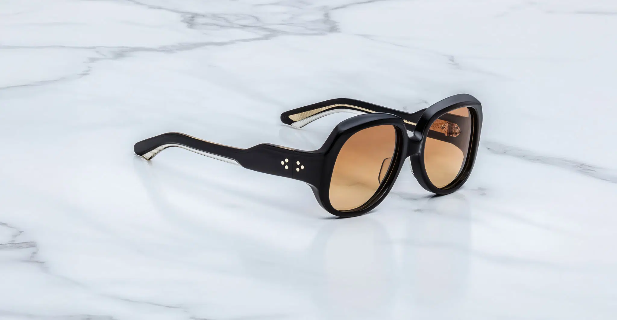 Jacques-Marie-Mage-Ocean-Child-Beluga-JMMYK-7T-3-4-Dan-Deutsch The Jacques Marie Mage Ocean Child sunglasses, featuring black thick frames, brown gradient lenses, and silver dot accents near the hinges, rest on a white marble surface.