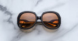 The Jacques Marie Mage Ocean Child, large round sunglasses with dark frames and amber-tinted lenses, are folded on a white marble surface with gray veining.