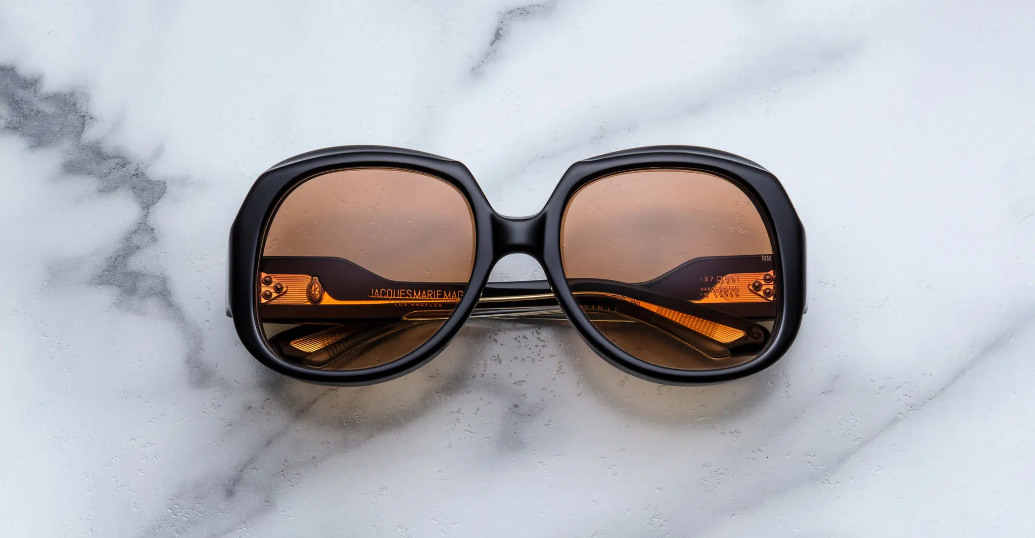 Jacques-Marie-Mage-Ocean-Child-Beluga-JMMYK-7T-Dan-Deutsch The Jacques Marie Mage Ocean Child, large round sunglasses with dark frames and amber-tinted lenses, are folded on a white marble surface with gray veining.