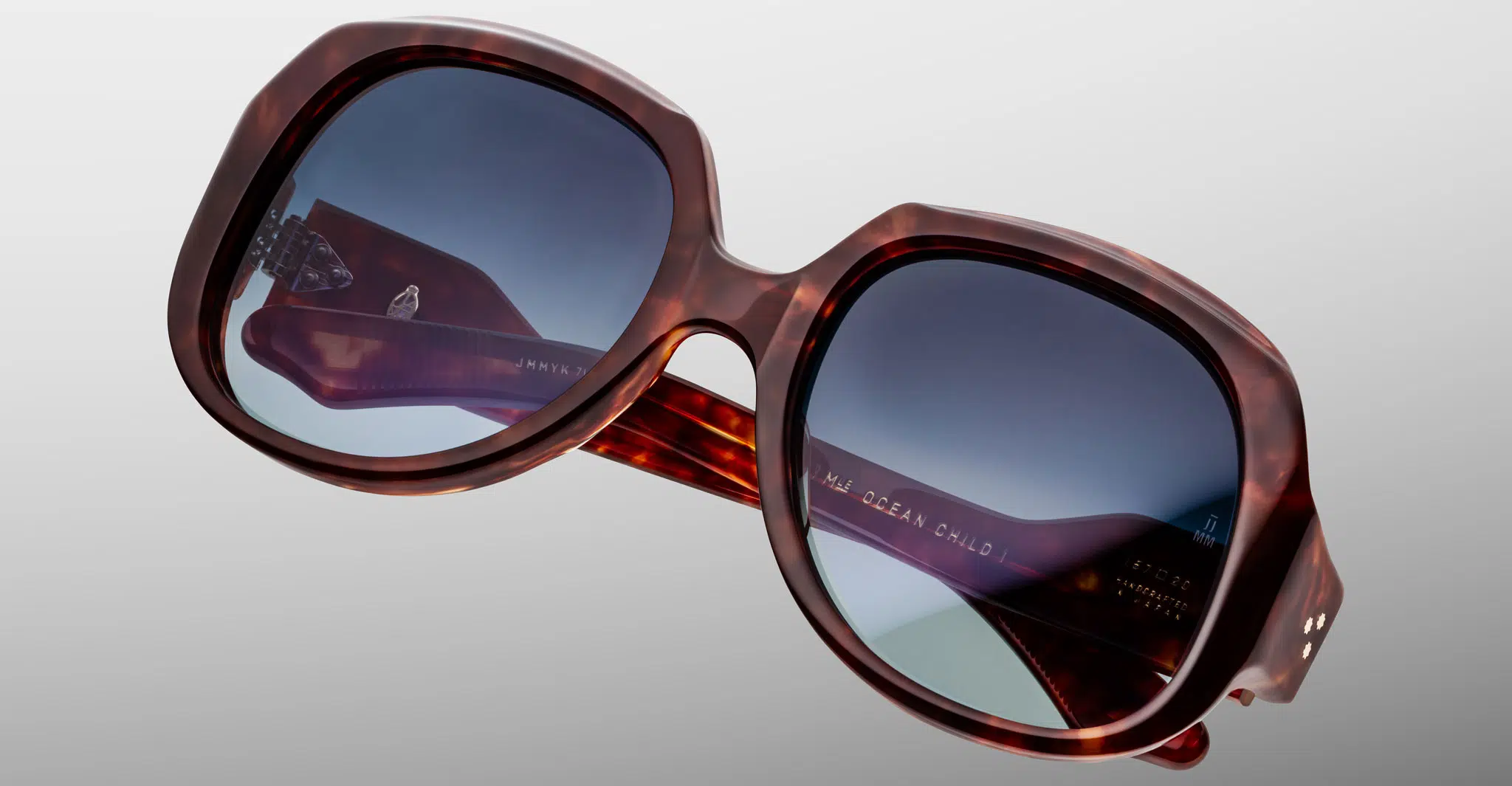 Close-up of oversized, square-shaped Jacques Marie Mage Ocean Child sunglasses in tortoiseshell with blue-tinted lenses, featuring engraved arm text reflecting light, set against a smooth light gray gradient background.