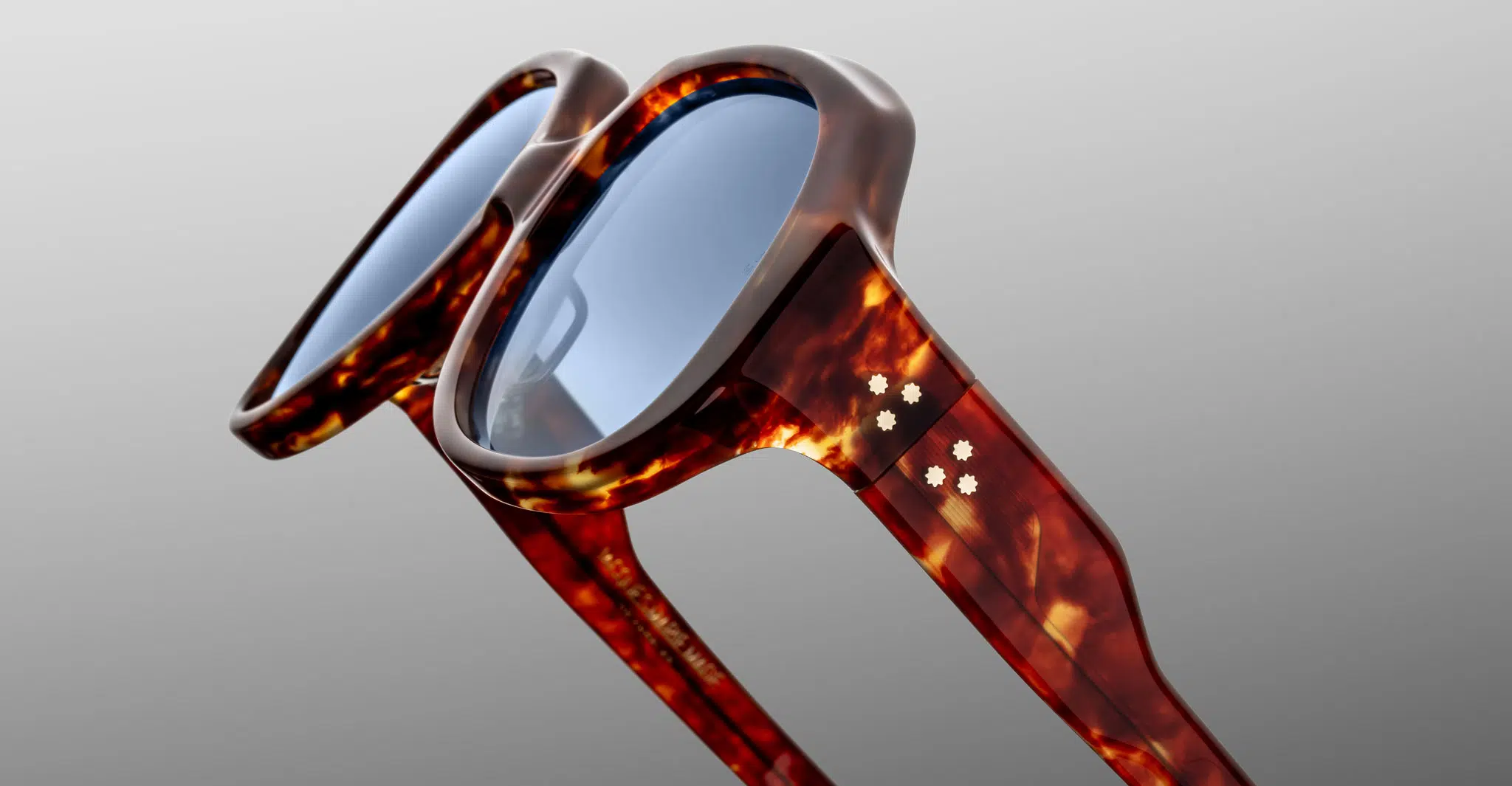 Close-up of Jacques Marie Mage Ocean Child sunglasses with stylish tortoiseshell frames, blue-tinted lenses, and decorative white star accents on the arms, set against a light gray background.