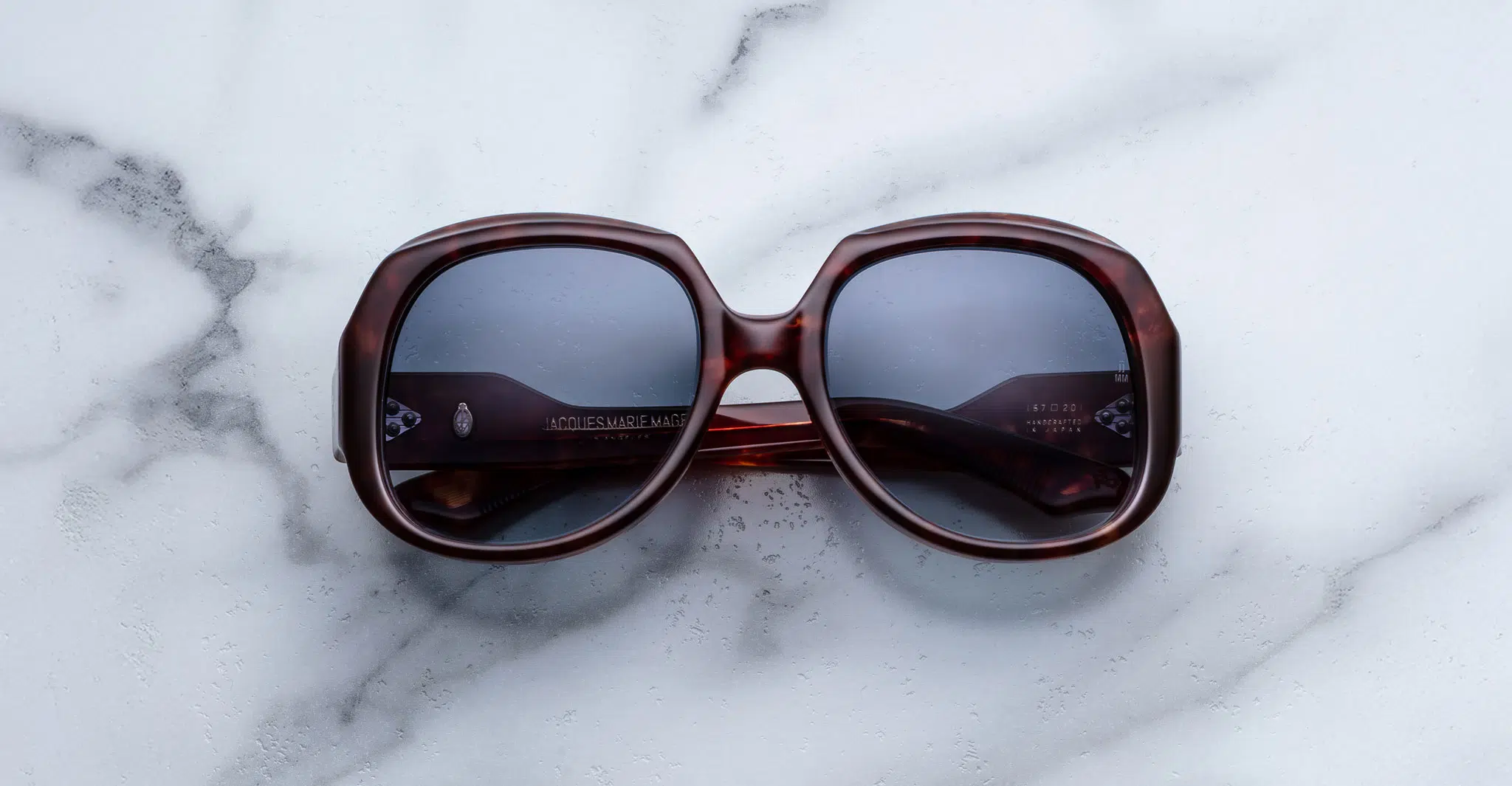 The Jacques Marie Mage Ocean Child sunglasses, featuring an oversized square design in dark brown with tinted lenses, rest on a white marble surface.