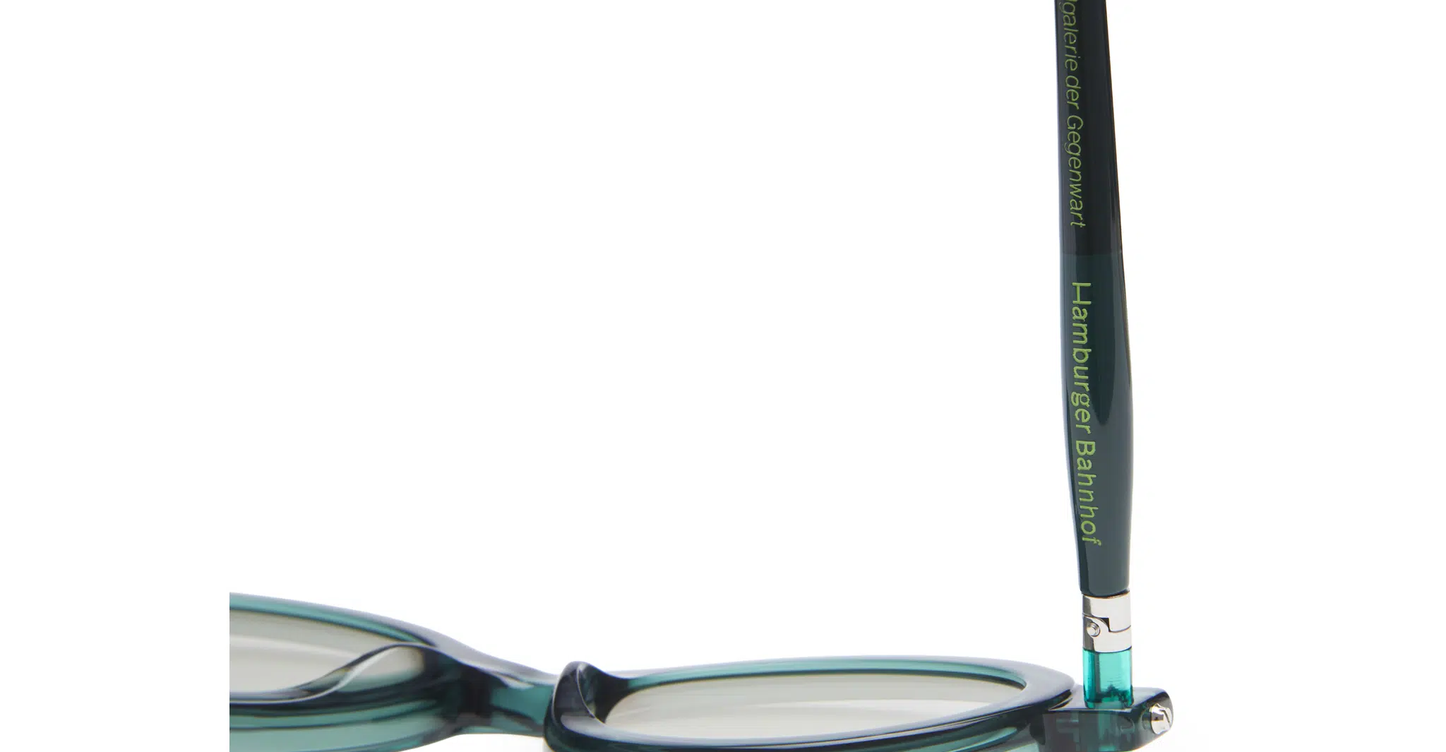 Close-up of green eyeglass frames with one temple arm raised. The temple arm has text in light green that reads Hamburger Bahnhof. The background is white.