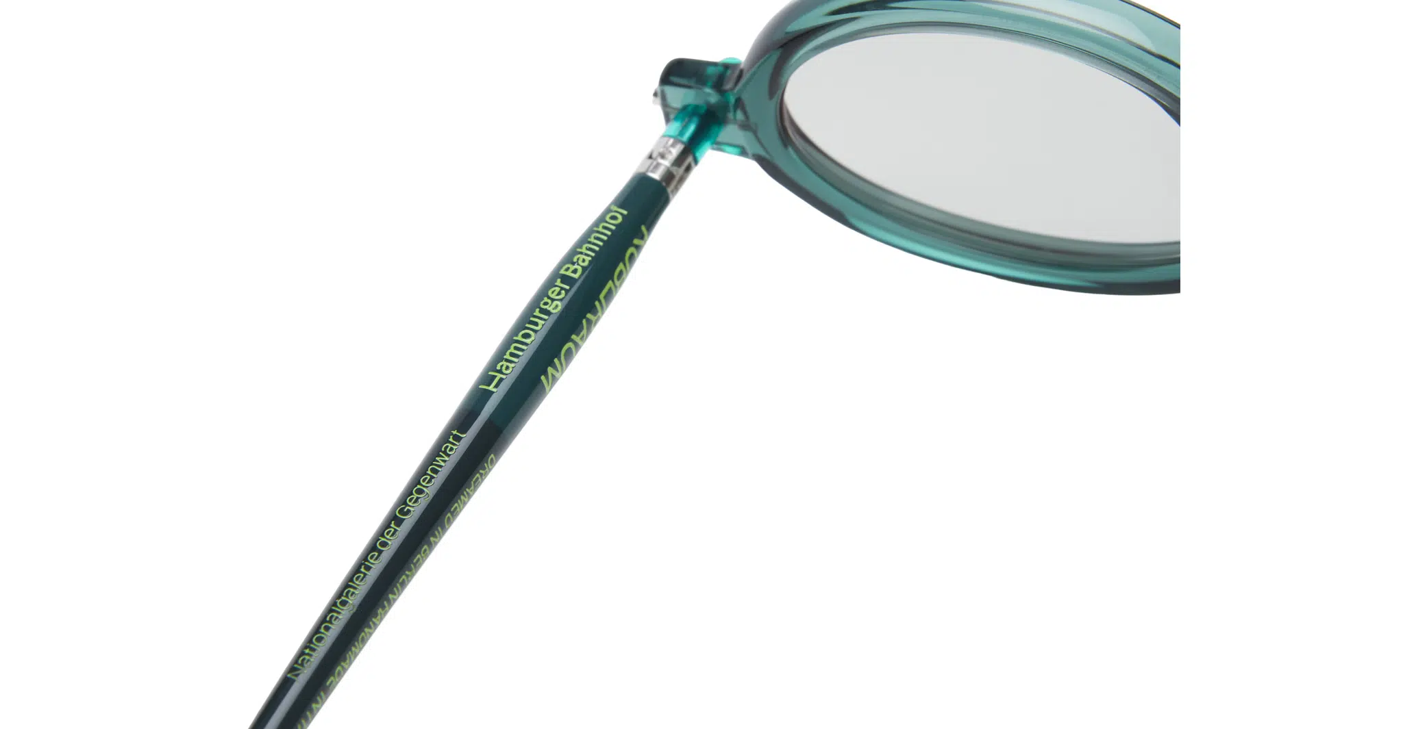 Close-up of the teal-colored arm of a pair of eyeglasses, with the words Hamburger Bahnhof and other text printed in yellow-green along the side. The clear lens and part of the round frame are visible.