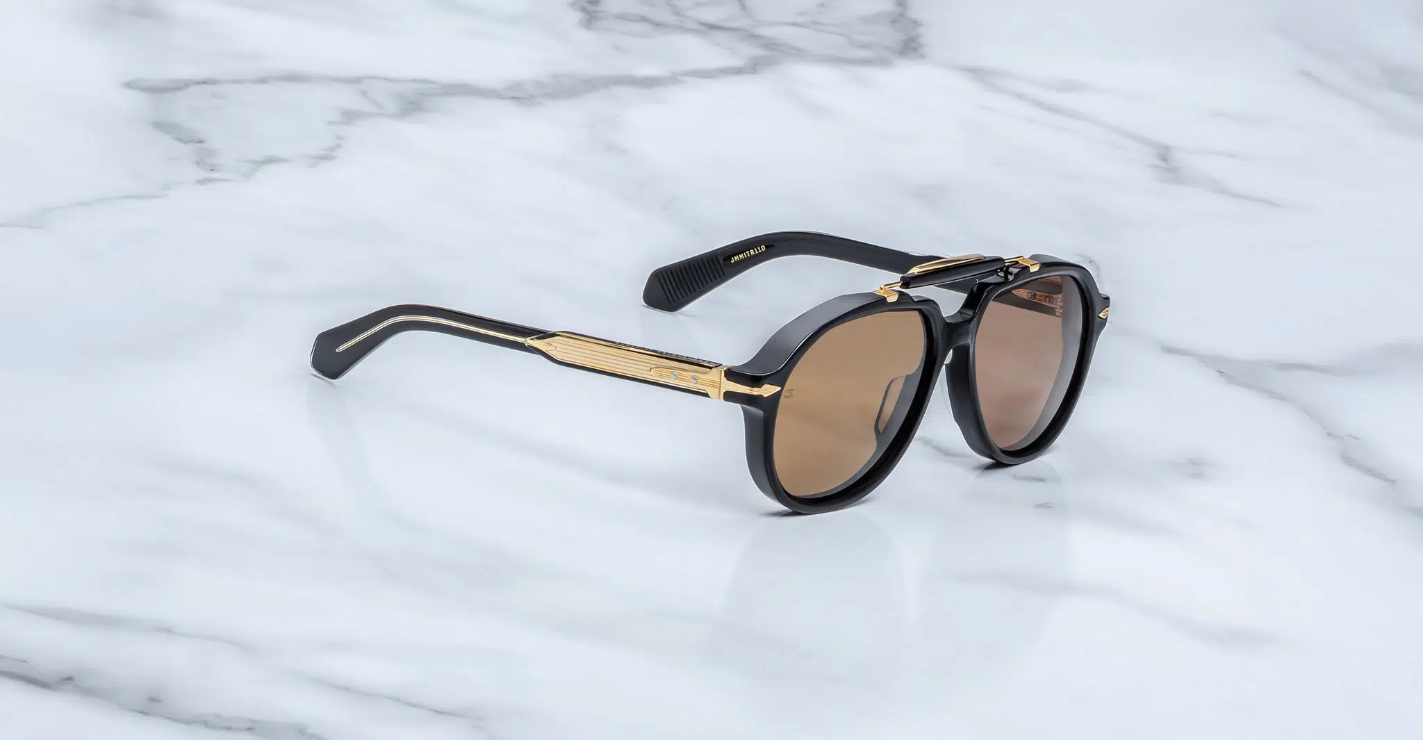 Jacques-Marie-Mage-1991-Eclipse-2-JMMITR-11D-3-4-Dan-Deutsch A pair of Jacques Marie Mage 1991 sunglasses with black frames, gold accents, and brown lenses rests on a white marble surface.