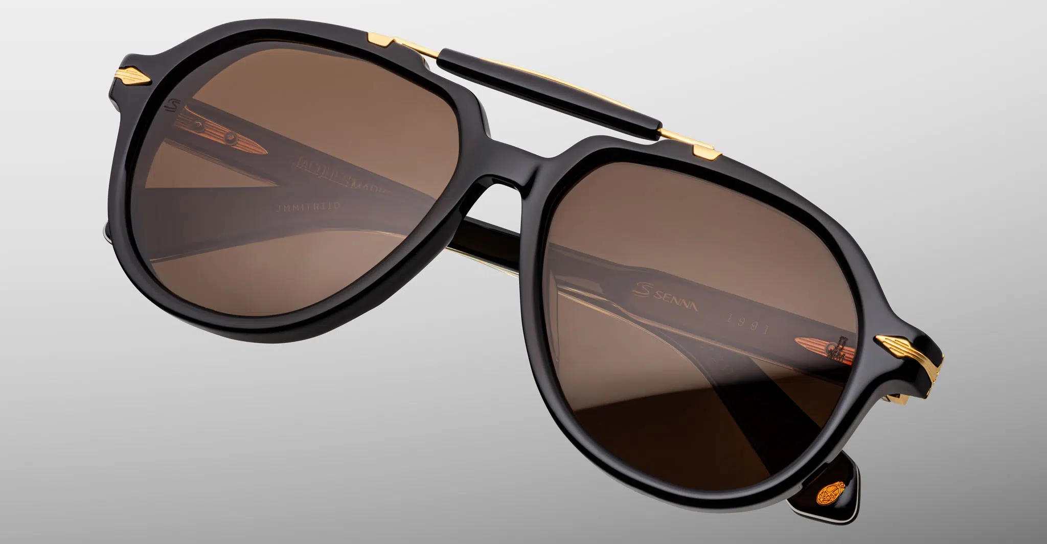 Jacques-Marie-Mage-1991-Eclipse-2-JMMITR-11D-Detail1-Dan-Deutsch The Jacques Marie Mage 1991 sunglasses feature a modern, angular design with black frames, brown lenses, and gold accents on the temples and bridge, displayed against a light gray background.