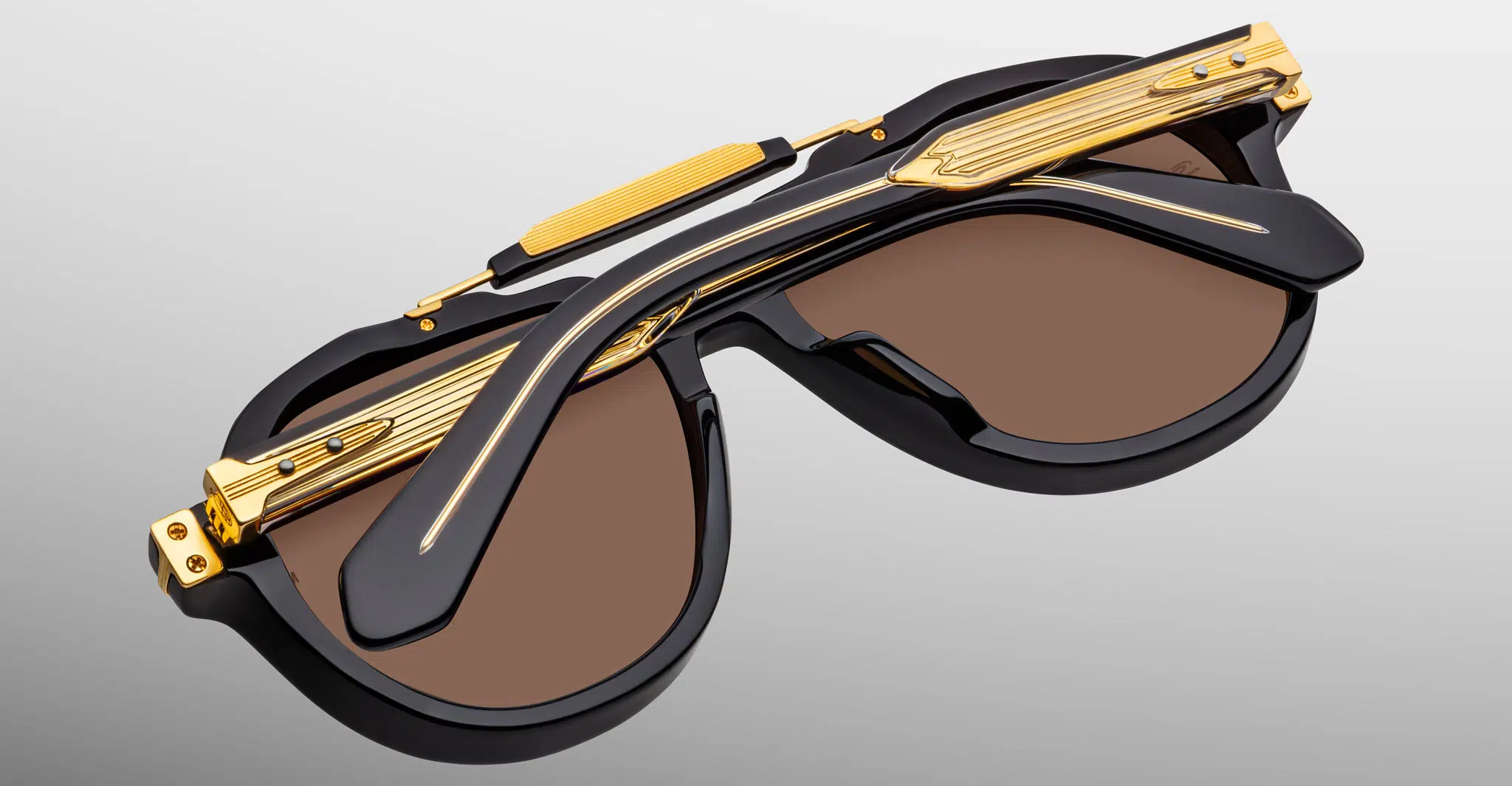 Jacques-Marie-Mage-1991-Eclipse-2-JMMITR-11D-Detail2-Dan-Deutsch A close-up of Jacques Marie Mage 1991 sunglasses with thick black frames, brown lenses, and decorative gold metal details on the arms and bridge, set against a light gray background.