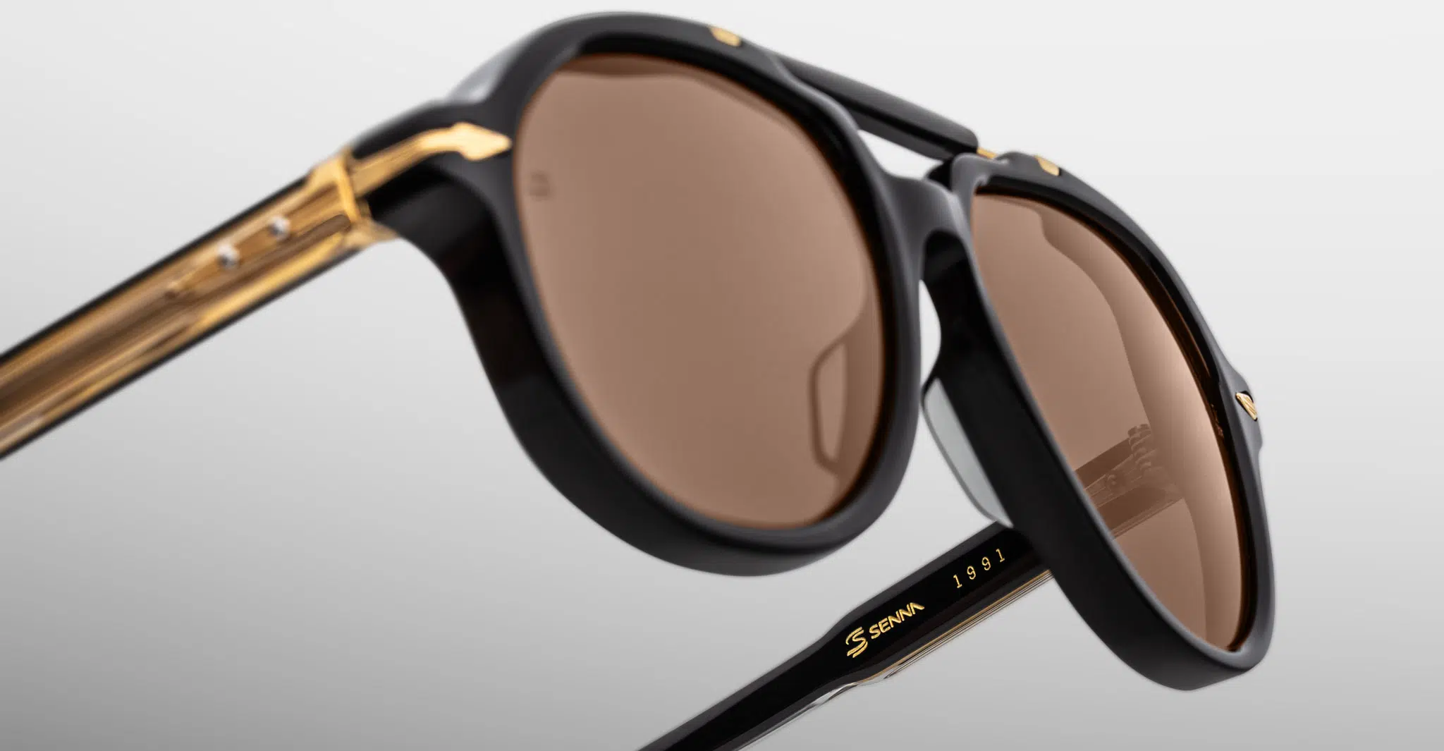 Jacques-Marie-Mage-1991-Eclipse-2-JMMITR-11D-Detail4-Dan-Deutsch Close-up of stylish black Jacques Marie Mage 1991 sunglasses with gold accents and brown-tinted lenses, featuring the brand name on the arm, set against a plain light background.