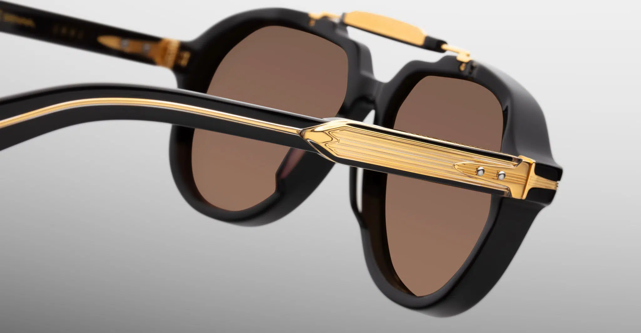 Jacques-Marie-Mage-1991-Eclipse-2-JMMITR-11D-Detail5-Dan-Deutsch Close-up of Jacques Marie Mage 1991 sunglasses in black with gold detailing and brown lenses, highlighting the intricate design on the arms and frame against a light gray background.