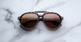 A pair of Jacques Marie Mage 1991 black sunglasses with brown-tinted lenses rests on a white marble surface, reflecting a modern railing and soft light in the lenses.