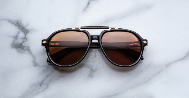 A pair of Jacques Marie Mage 1991 black sunglasses with brown-tinted lenses rests on a white marble surface, reflecting a modern railing and soft light in the lenses.