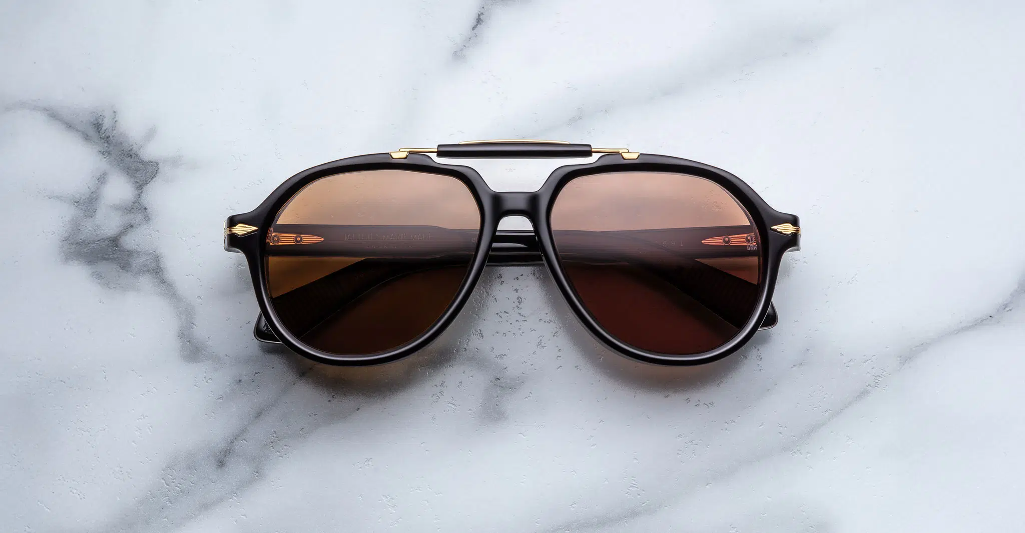 Jacques-Marie-Mage-1991-Eclipse-2-JMMITR-11D-Front-Dan-Deutsch A pair of Jacques Marie Mage 1991 black sunglasses with brown-tinted lenses rests on a white marble surface, reflecting a modern railing and soft light in the lenses.