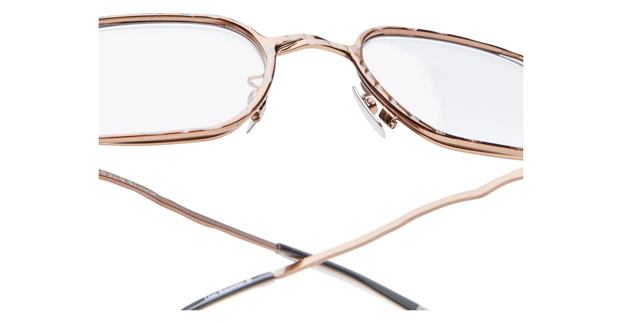 Kuboraum-H22-BZ-Bronze-Optical-KROH22BZ000000OP-Detail1-Dan-Deutsch Close-up view of Kuboraum H22 eyeglasses in rose gold metal with clear lenses, adjustable nose pads, and partially visible temple arms crossed at the bottom against a white background.