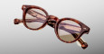 The Jacques Marie Mage Clemens features round, brown tortoiseshell eyeglasses with thick frames and gold temple detailing, shown against a light gray background.