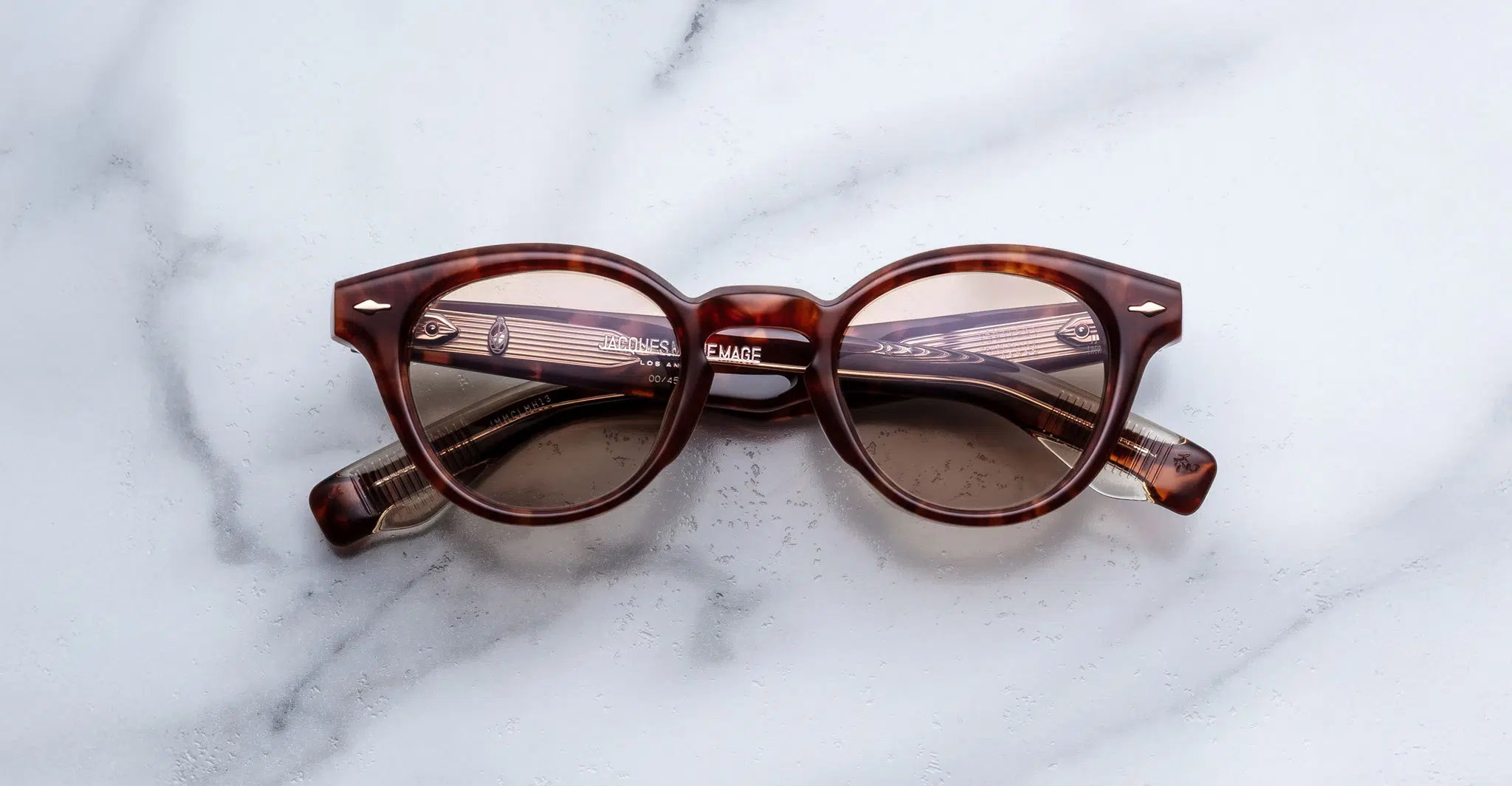 Jacques Marie Mage Clemens round tortoiseshell eyeglasses with clear lenses are placed upside down on a white marble surface, their temples crossing over each other.