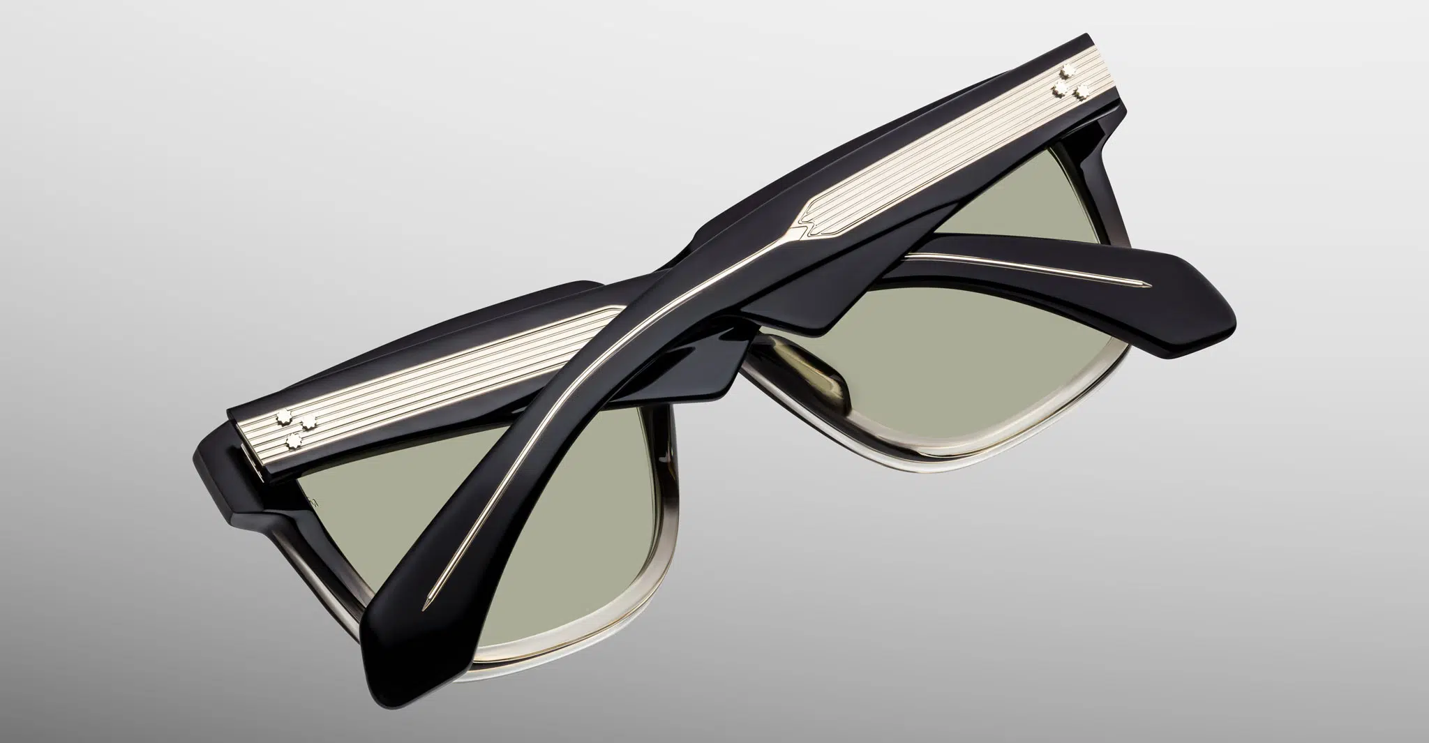 The Jacques Marie Mage Hyannis sunglasses feature thick black frames, gold accents, and light green lenses, shown folded on a light gray background.