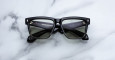 A pair of Jacques Marie Mage Hyannis black thick-framed rectangular sunglasses with dark lenses is displayed on a white marble surface, arms folded to reveal the inside branding.