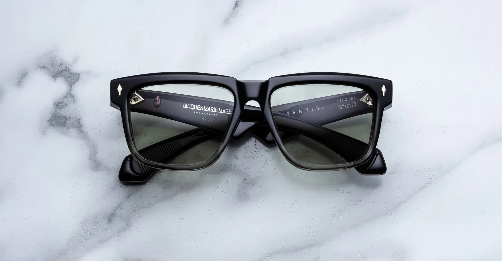 A pair of Jacques Marie Mage Hyannis black thick-framed rectangular sunglasses with dark lenses is displayed on a white marble surface, arms folded to reveal the inside branding.