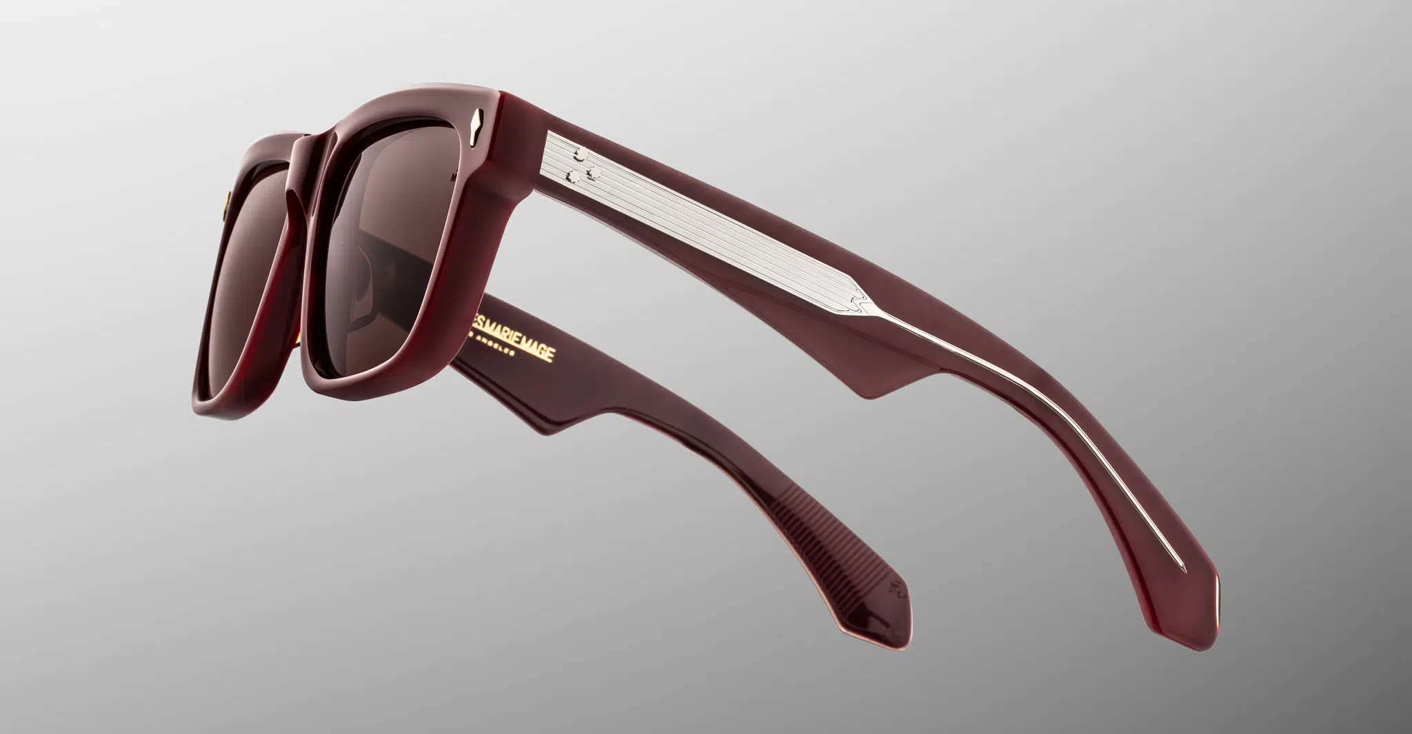 The Jacques Marie Mage Hyannis sunglasses feature thick dark red frames, metallic arm accents, and tinted lenses, showcased on a light gray gradient background.