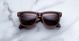A pair of Jacques Marie Mage Hyannis dark brown square sunglasses with dark lenses rests on a white marble surface.