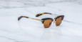 A pair of Jacques Marie Mage Sartre sunglasses with brown lenses, gold accents, and black and tortoiseshell-patterned frames rests on a white marble surface.