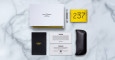 A minimalist flat lay on marble showcases the Jacques Marie Mage Torrance in a black case, a white box, silver pen, yellow cloth with 237 in bold black, a black notebook, and two cards.