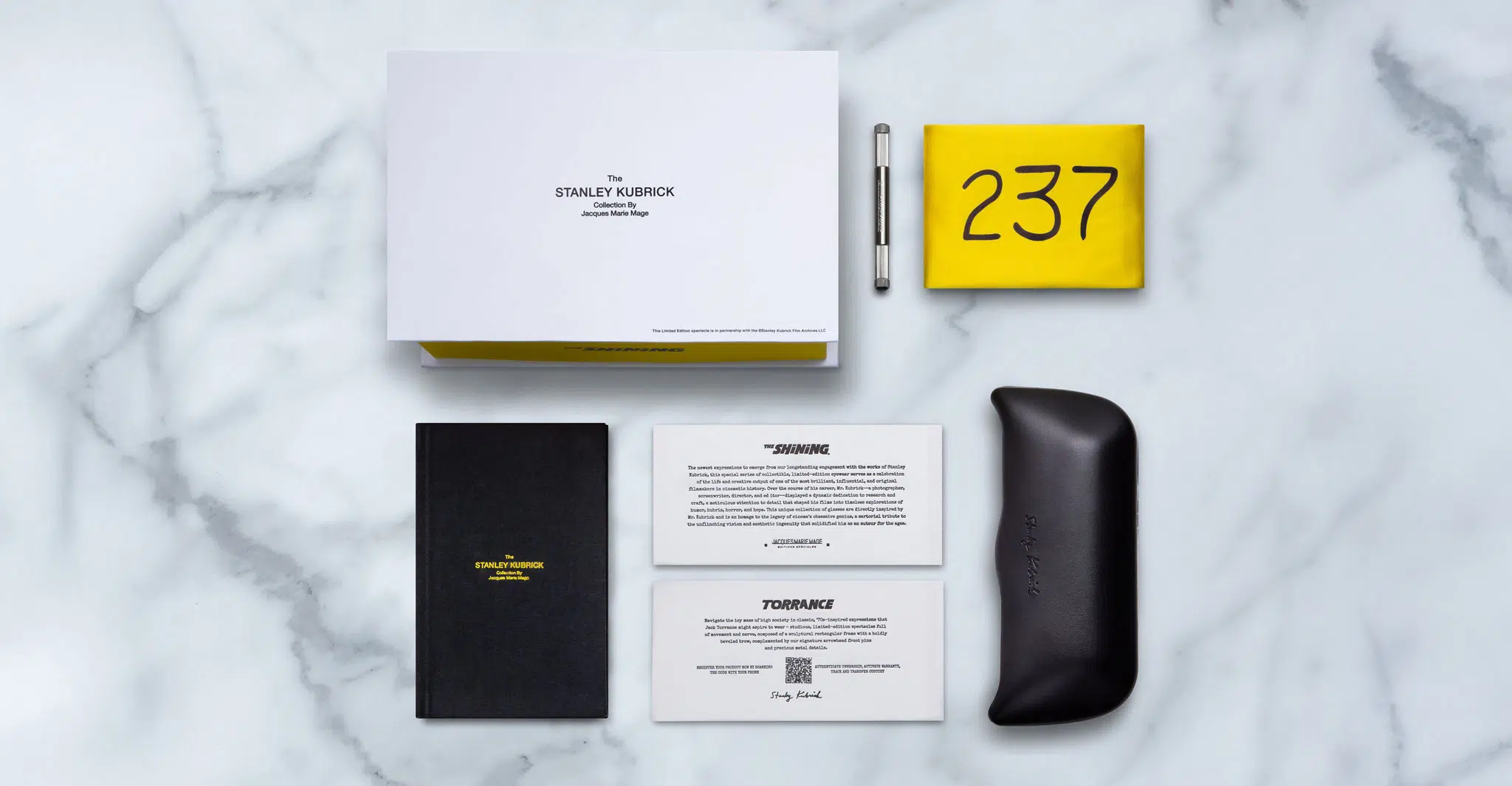 A minimalist flat lay on marble showcases the Jacques Marie Mage Torrance in a black case, a white box, silver pen, yellow cloth with 237 in bold black, a black notebook, and two cards.