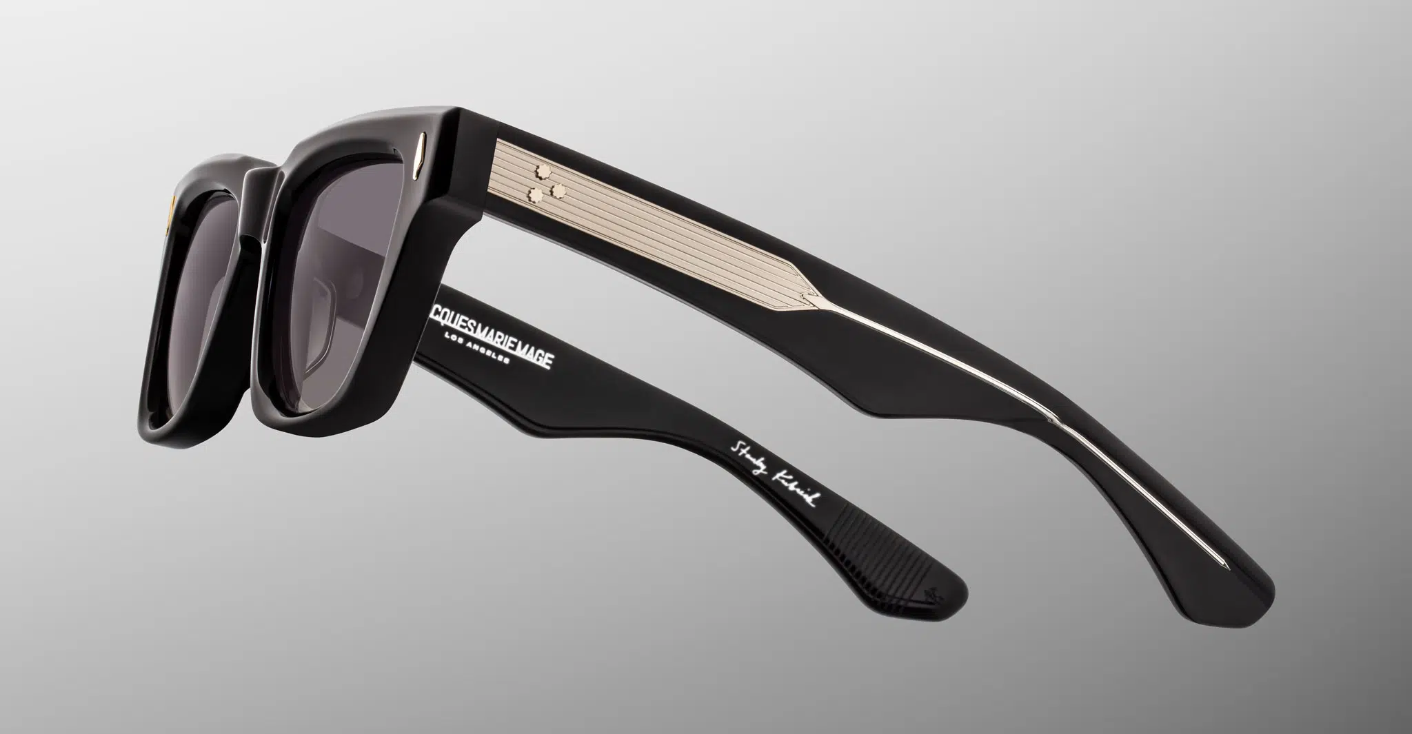 Jacques Marie Mage Torrance sunglasses, featuring dark lenses, gold-accented arms, and white branding on the inner temple, are shown against a gradient gray background.
