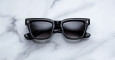 A pair of Jacques Marie Mage Torrance black rectangular sunglasses with dark lenses rests on a white marble surface with grey veining.
