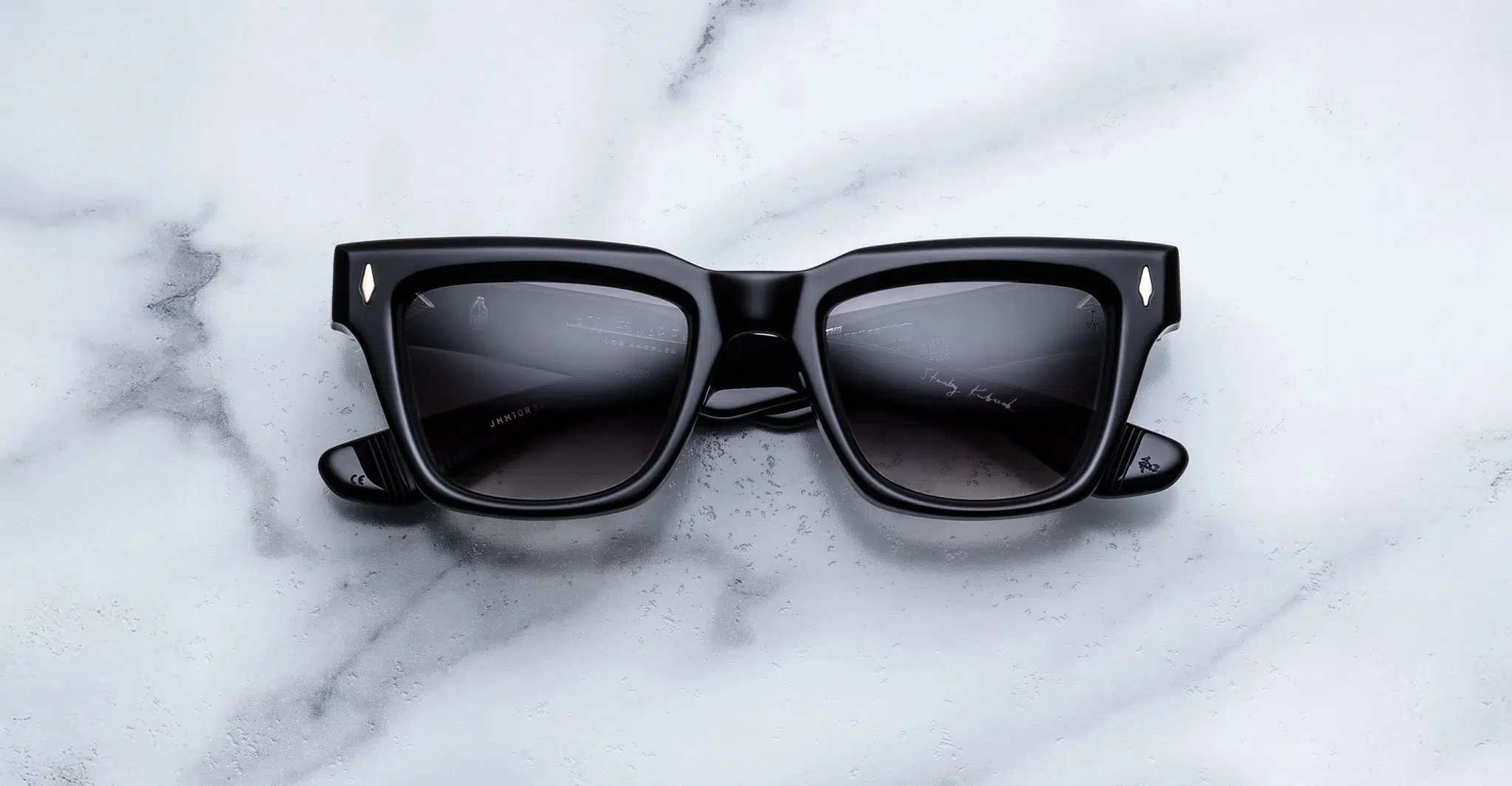 A pair of Jacques Marie Mage Torrance black rectangular sunglasses with dark lenses rests on a white marble surface with grey veining.