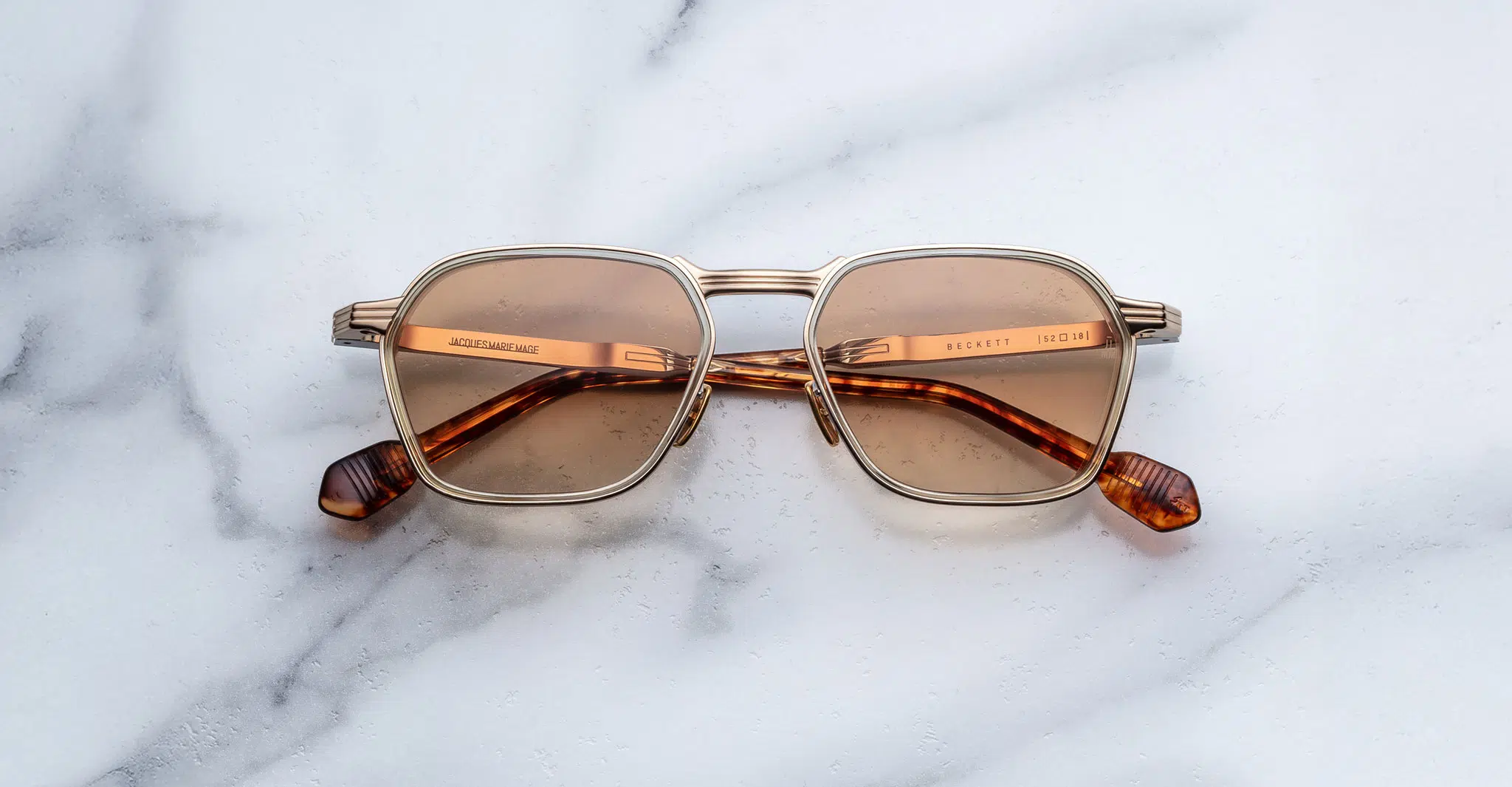 The Jacques Marie Mage Beckett sunglasses, featuring brown lenses and gold and tortoiseshell frames, rest on a white marble surface.